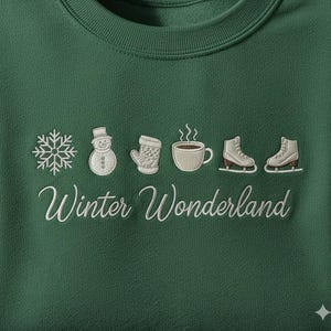Op de afbeelding: Een donkergroene sweatshirt met witte geborduurde winterontwerpen. Het ontwerp bevat een sneeuwvlok, sneeuwpop, want, koffiekopje en schaatsen, met de woorden "Winter Wonderland" in cursief.