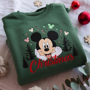 May include: A green sweatshirt featuring an embroidered Mickey Mouse design with the word "Christmas" in red. The design includes Christmas trees, candy canes, and Mickey Mouse head shapes. The sweatshirt is folded.