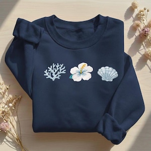 Op de afbeelding: Marineblauwe sweatshirt met geborduurd koraal, een witte hibiscusbloem en een zeeschelp. De sweatshirt is op een licht houten oppervlak gevouwen. De borduursels zijn in tinten blauw, wit en geel.