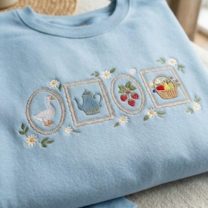 Könnte beinhalten: Hellblaues Sweatshirt mit gestickten, gerahmten Bildern einer Gans, einer Teekanne, Erdbeeren und einem Obstkorb. Die Stickerei ist in Blau-, Grün- und Rottönen gehalten, mit weißen Gänseblümchen-Akzenten. Das Sweatshirt ist aus weichem Material.