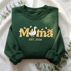Può includere: Una felpa verde scuro con la scritta "Momma" in lettere ricamate gialle, con un disegno di anatra e anatroccolo. Il testo "EST. 2026" è sotto la parola "Momma". Farfalle e fiori sono ricamati anche sulla felpa.