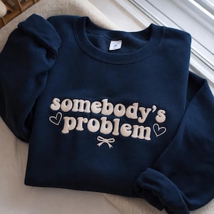 Pode incluir: Camisola azul marinho com a frase bordada "somebody's problem" em uma fonte cremosa e divertida. O design inclui detalhes de coração e laço. A camisola é feita de um material macio e tem um decote redondo clássico.