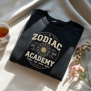 May include: Black sweatshirt with a gold and cream-colored graphic. The graphic reads "ZODIAC ACADEMY" with a sun and zodiac symbols. The sweatshirt also has the text "TERRA AER IGNIS AQUA" and the year "2018".