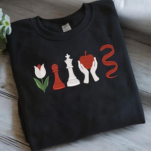 May include: Black crewneck sweatshirt with embroidered design. The design features a white and red tulip, a red pawn, a white king chess piece, a red apple held by white hands, and a red ribbon.