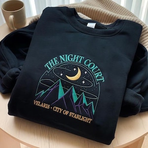 Könnte beinhalten: Schwarzes Sweatshirt mit einem gestickten Design mit einem Halbmond, Sternen und Bergen. Der Text "THE NIGHT COURT" und "VELARIS • CITY OF STARLIGHT" ist ebenfalls auf dem Sweatshirt gestickt.