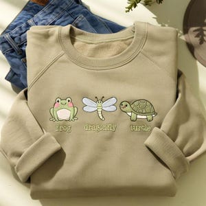 以下が含まれることがあります： セージグリーンのスウェットシャツ。カエルの刺繍、トンボ、カメのデザイン。各動物の下には「frog」、「dragonfly」、「turtle」の文字が刺繍されています。背景には青いジーンズが折りたたまれています。