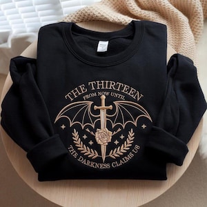 May include: Black sweatshirt with gold embroidery. The design features the text "THE THIRTEEN" above a sword and wing emblem, with "FROM NOW UNTIL" and "THE DARKNESS CLAIMS US" below. A rose and laurel wreath are also included.