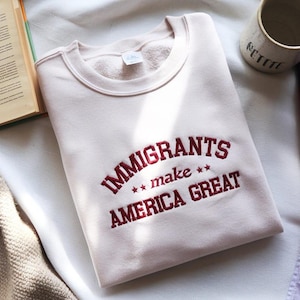 Puede incluir: Sudadera rosa claro con la frase bordada en borgoña "IMMIGRANTS make AMERICA GREAT". La sudadera tiene cuello redondo y mangas largas. La imagen está tomada desde arriba.