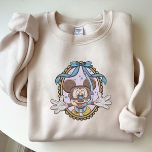 May include: A beige sweatshirt featuring an embroidered design of Mickey Mouse wearing a snorkel and mask. The design is framed by a light blue bow and a scalloped border. The sleeves are folded.