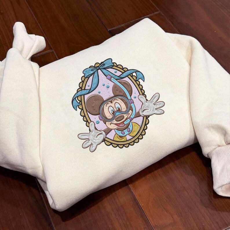 May include: Cream-colored sweatshirt featuring an embroidered design of Mickey Mouse wearing a snorkel and diving mask. The design is framed in a scalloped oval with a blue bow at the top. The sweatshirt has ribbed cuffs and a folded hem.