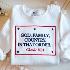 Może przedstawiać: Biała bluza z prostokątnym wzorem w kolorze czerwonym i niebieskim. Wzór zawiera tekst "GOD, FAMILY, COUNTRY. IN THAT ORDER. Charlie Kirk" z gwiazdkami. Swobodny, wygodny top.