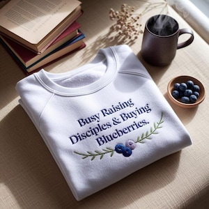 May include: White sweatshirt with the embroidered text "Busy Raising Disciples & Buying Blueberries." The design includes a blueberry and leaf motif. A mug of coffee, a small bowl of blueberries, and books are in the background.