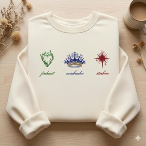 Peut inclure: Sweat-shirt crème avec trois motifs brodés : un cœur vert avec le mot "fireheart", une couronne bleue et dorée avec "cursebreaker", et une étoile rouge avec "starborn". Les manches sont retroussées.