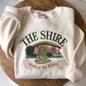 May include: Cream-colored sweatshirt with the embroidered text "THE SHIRE" above a green and brown illustration of a hobbit hole. The banner below reads "Home of the Hobbits". The sleeves are cuffed.