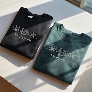 May include: Two crewneck sweatshirts, one black and one dark green, with white embroidered designs. Each features a seashell, turtle, and starfish, with the text "Be still and know that I am God, Psalm 46:10."