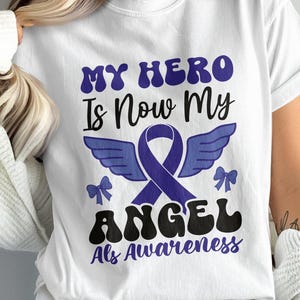 May include: White t-shirt with the text "My Hero Is Now My Angel ALS Awareness" in black and purple lettering. The text is in a retro font style. A purple ribbon with wings is behind the text.