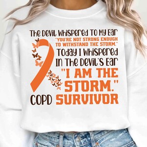 Puede incluir: Sudadera blanca con una cinta naranja y mariposas. El texto dice: "El diablo me susurró al oído... Hoy susurré al oído del diablo: 'Yo soy la tormenta.' Superviviente de EPOC."