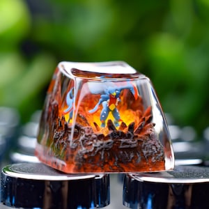 Puede incluir: Una tecla rectangular transparente con una escena en miniatura detallada en su interior. La escena representa una figura azul en un entorno de fuego, naranja y marrón. La tecla está colocada en un teclado.