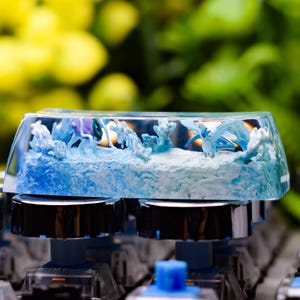 May include: A custom keyboard keycap with a clear resin top, featuring a detailed ocean scene with blue and white waves and a dragon figure. The keycap sits on a black and silver keyboard.