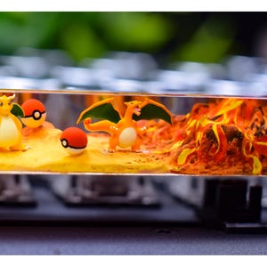 May include: A custom keyboard keycap with a miniature diorama. It features Pokémon characters Charizard and Dragonite, two Poké Balls, and a fiery volcanic landscape. The keycap is transparent, showcasing the detailed scene within.