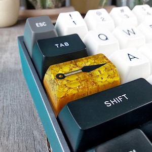 Peut inclure: Un clavier mécanique bleu-vert avec un capuchon de touche jaune et or représentant une étoile noire de lancer. Le capuchon de touche est étiqueté "SHIFT".