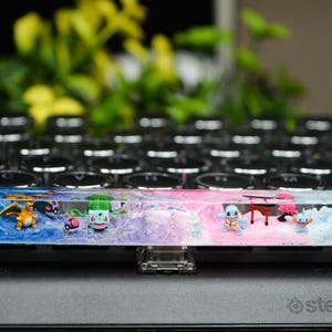 May include: A custom keyboard spacebar with a resin design featuring various Pokémon characters. The spacebar has a blue, pink, and purple colour scheme, with small figurines of Pokémon and a miniature table. The keyboard is black.