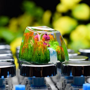May include: A custom keycap with a miniature diorama showing a waterfall, green grass, and orange flowers. A pink cartoon character is visible inside the clear resin keycap, which sits on a mechanical keyboard.
