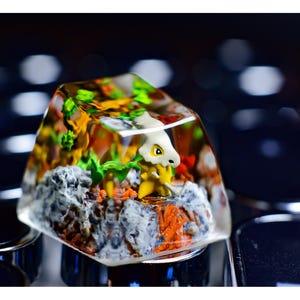 Puede incluir: Un tapón de tecla de resina transparente con una pequeña figura de dinosaurio amarillo y blanco en su interior. El dinosaurio está rodeado de resina verde y naranja, que se asemeja a un paisaje en miniatura.