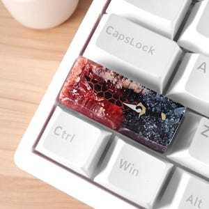 Puede incluir: Un tapón de tecla de resina rojo y negro con un patrón de panal y un diseño estilizado. El tapón de tecla está en un teclado blanco con teclas etiquetadas "Tab", "CapsLock", "Ctrl", "Win", "Alt" y "A".