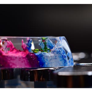 Peut inclure: Un capuchon de touche en résine transparente avec un motif abstrait rose, bleu et vert. Le motif présente une petite figurine verte au centre.
