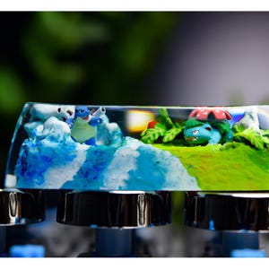 Peut inclure: Un keycap personnalisé avec un diorama miniature de personnages Pokémon. Le keycap a un dessus en résine transparente, montrant une scène avec de l'eau bleue, de l'herbe verte et des figurines Pokémon. Le keycap est monté sur un clavier.