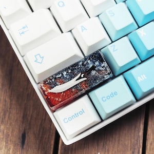 Puede incluir: Un teclado blanco con teclas azules y azul claro. La tecla 'Control' tiene una tecla personalizada con un diseño de resina de una espada en un paisaje nevado.