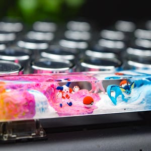May include: A custom keyboard spacebar with a colorful, abstract design. The spacebar features miniature figurines of cartoon characters in a clear resin, with a mix of pink, blue, yellow, and orange hues.