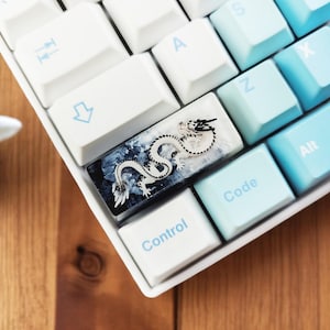 Puede incluir: Un teclado blanco con una tecla personalizada que presenta un diseño de dragón blanco y negro. La tecla está hecha de resina transparente y tiene un degradado azul y blanco.