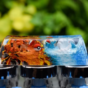 May include: A custom keycap with a split design, showing a fiery orange landscape and a blue water scene. The keycap features miniature cartoon characters and a red and white ball. The keycap is mounted on a mechanical keyboard.