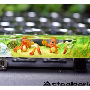 May include: A custom keycap featuring two orange Charmander figures in a miniature landscape with green and yellow terrain. The keycap is set against a keyboard background, with a clear resin top.
