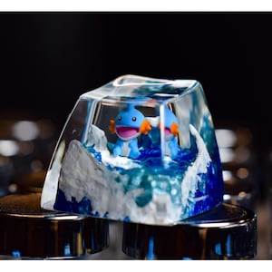 May include: A clear resin keycap featuring a blue and white ocean scene. Two small blue and orange figurines are inside the resin, resembling aquatic creatures. The keycap is resting on a black surface.