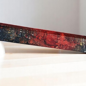 Può includere: Un oggetto rettangolare multicolore con un motivo rosso, nero e oro, appoggiato su una base geometrica bianca. L'oggetto sembra essere fatto di resina o materiale simile. Il design generale è moderno e artistico.