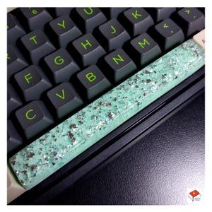 May include: A custom keyboard space bar with a mint green resin base and silver glitter accents. The space bar is resting on a black keyboard with dark gray keys and green lettering.