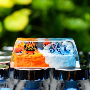 Peut inclure: Une touche de clavier personnalisée avec un design divisé en sections orange et bleue. Le côté orange présente un personnage Pokémon et une Poké Ball, le côté bleu un autre personnage Pokémon et une Poké Ball. La touche est posée sur un clavier.