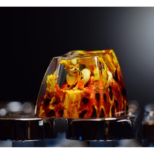 May include: A clear resin keycap with a small orange and yellow figurine of a fox inside. The resin is swirled with orange and yellow, resembling flames. The keycap is sitting on a black keyboard.