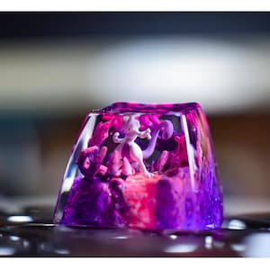 Può includere: Un copritasto in resina trasparente con un design a spirale rosa e viola. Una piccola statuina bianca di un personaggio Pokemon è all'interno del copritasto.