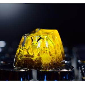 Puede incluir: Un tapón de tecla de resina amarilla con una pequeña criatura amarilla en su interior. El tapón de tecla está sobre un teclado negro.