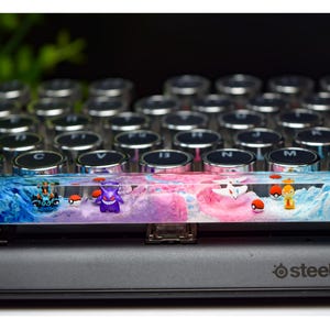 Puede incluir: Un tapón de tecla de teclado de resina transparente con un diseño de remolino azul y rosa. El tapón de tecla presenta pequeñas figuras de personajes de Pokémon, incluyendo un Gengar, un Jigglypuff y un Pikachu.