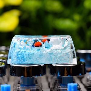 Peut inclure: Un keycap personnalisé représentant une scène océanique miniature avec des vagues bleues, des icebergs blancs et des personnages de dessins animés. Le keycap transparent est monté sur un clavier mécanique.