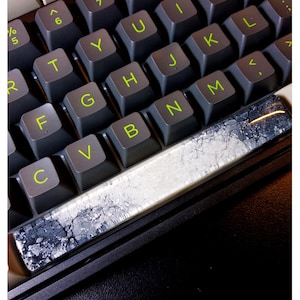 Puede incluir: Primer plano de un teclado mecánico con teclas grises y letras verde brillante. La barra espaciadora tiene un diseño único con un patrón veteado blanco y gris. El teclado está sobre una superficie negra.