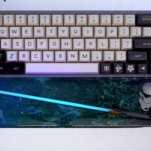 May include: A white mechanical keyboard with black keycaps and a blue and green galaxy-themed mouse mat. The mouse mat has a blue glow stick and a white Stormtrooper figurine.
