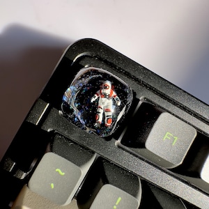 May include: A white and red astronaut figurine is encased in a clear resin keycap with a black and blue galaxy background. The keycap is on a black keyboard with a grey F1 key.