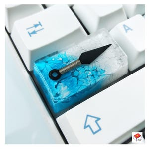 May include: A blue and white resin keycap with a black throwing star embedded in the resin. The keycap is on a white keyboard.