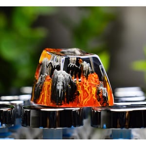May include: A custom keycap with a mountain and lava design. The keycap is clear with a detailed interior scene featuring black, white, and orange elements. The keycap sits atop a row of silver and black mechanical keyboard components.